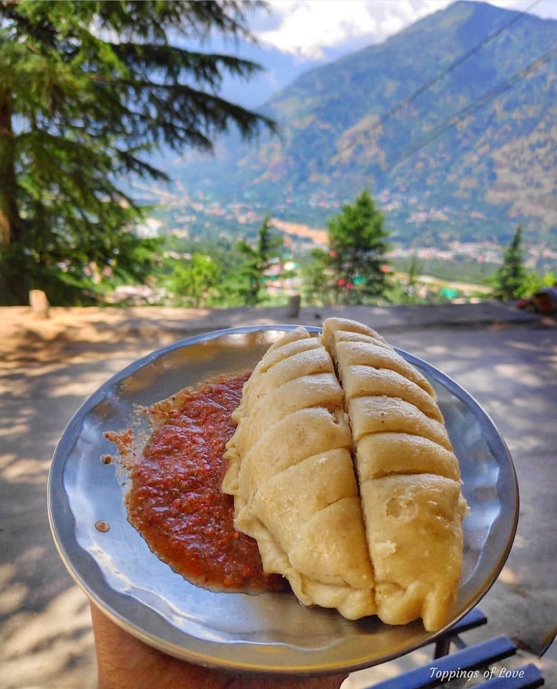 Sliced Siddu served with red garlic chutney, held up against a panoramic view of the Kullu valley